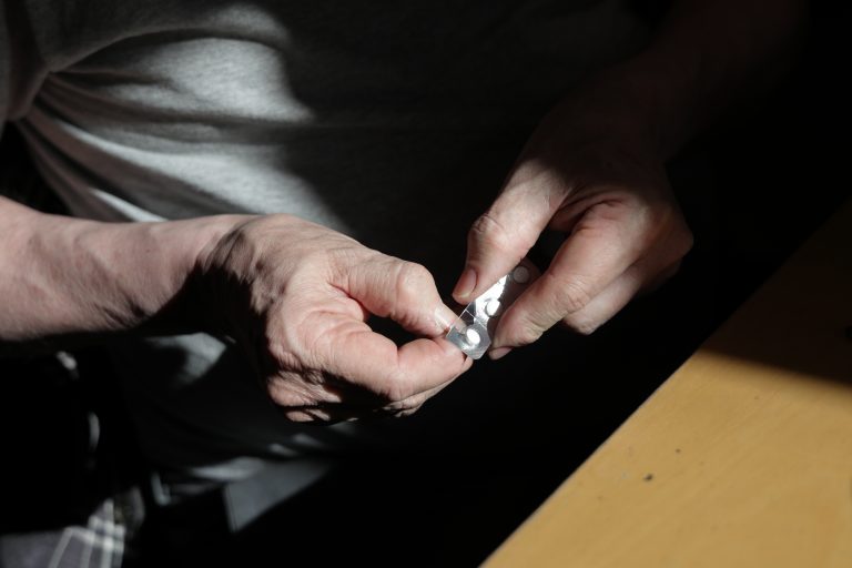 A senior Canadian man with prostate cancer opens a blister pack to release a dose of nonsteroidal antiandrogen. Bicalutamide. Ambient light inside a dining room in a British Columbia home.