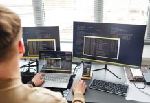 Rear view of young IT specialist connecting laptop with computer while typing data on keyboard sitting at his workplace, three screens showing the same thing, a virtual desktop, VDI