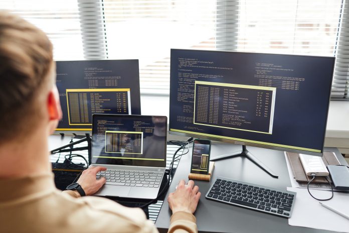 Rear view of young IT specialist connecting laptop with computer while typing data on keyboard sitting at his workplace, three screens showing the same thing, a virtual desktop, VDI