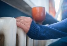 Man in warm sweater with hot tea near radiator in winter. Energy crisis concept.