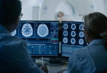 In Control Room Doctor and Radiologist Discuss Diagnosis while Watching Procedure and Monitors Showing Brain Scans Results, In the Background Patient Undergoes MRI or CT Scan Procedure.