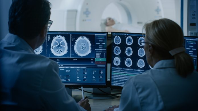 In Control Room Doctor and Radiologist Discuss Diagnosis while Watching Procedure and Monitors Showing Brain Scans Results, In the Background Patient Undergoes MRI or CT Scan Procedure.