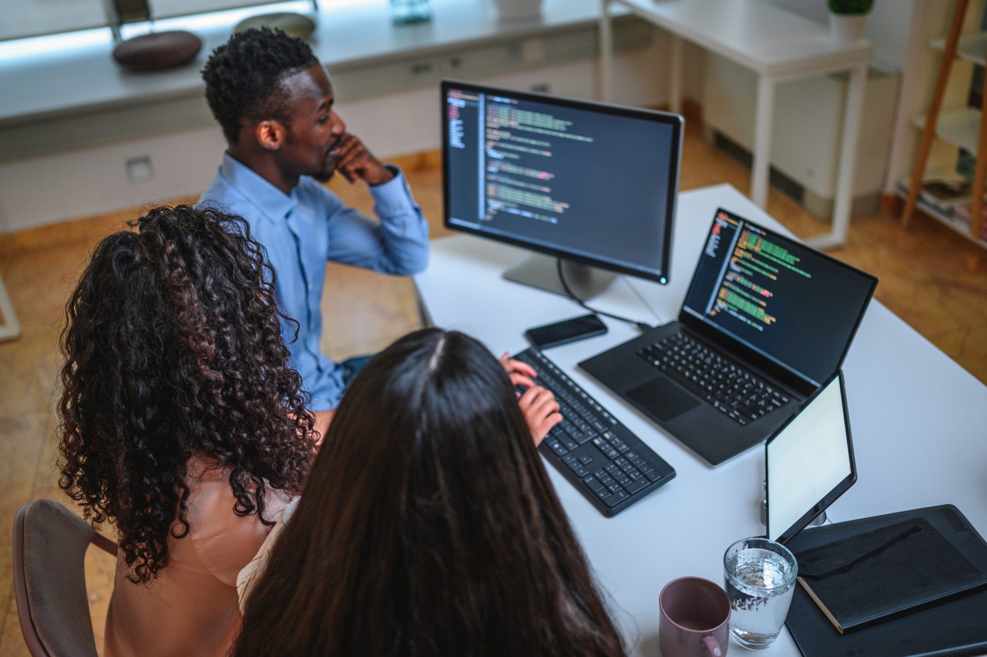 Black businessman, Asian businesswoman and Hispanic businesswoman are programming in an open plan office together. They are using computer, laptop and digital tablet.