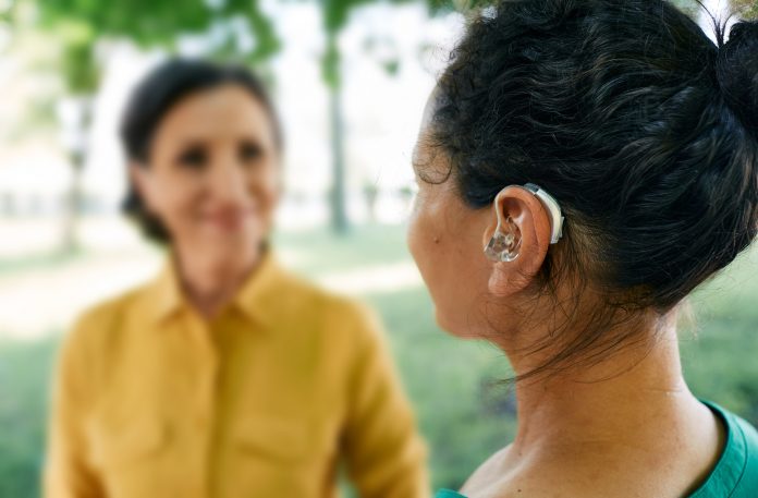 Adult woman with a hearing impairment uses a hearing aid to communicate with her female friend at city park. Hearing solutions, sensorineural hearing loss