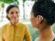 Adult woman with a hearing impairment uses a hearing aid to communicate with her female friend at city park. Hearing solutions, sensorineural hearing loss