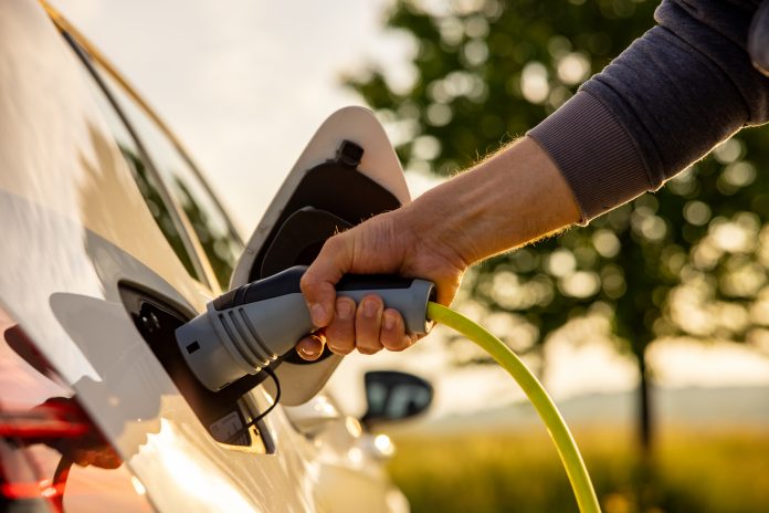 Hand of man inserting a power cord into an electric car for charging ecofriendly vehicle on green landscape