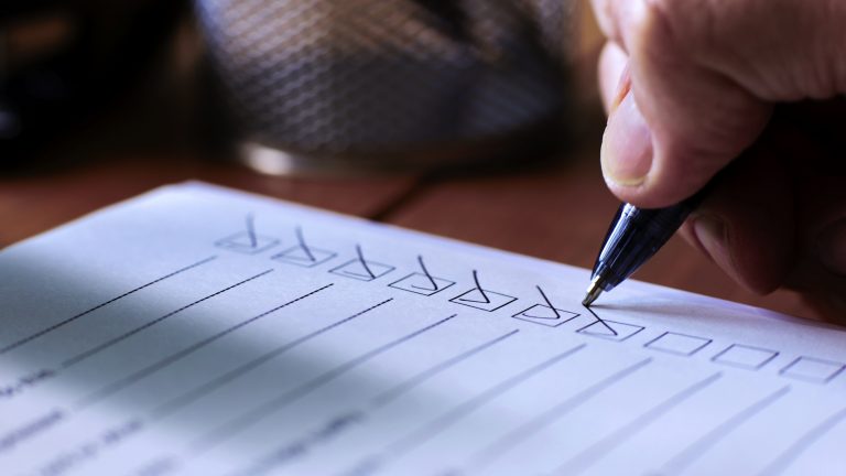 Close up image of a person’s hand using a ballpoint pen to put a check or tick mark in a row of boxes on a paper form sheet.