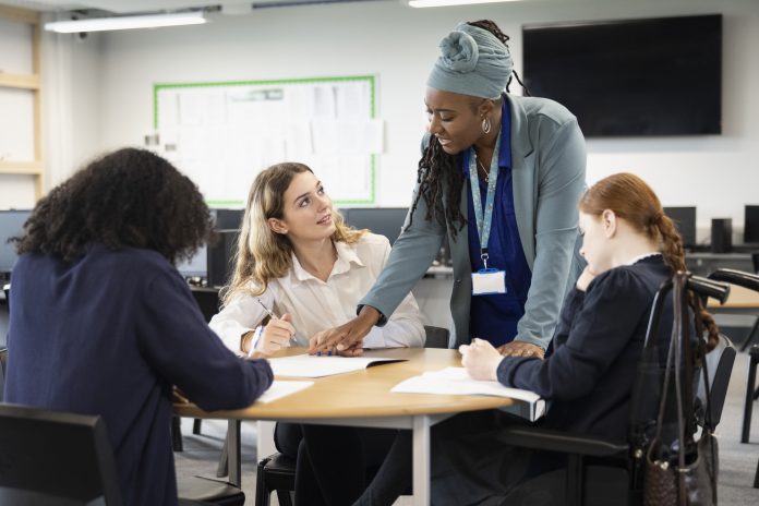 Waist-up view of 15 and 16 year old classmates in uniforms sitting at round table taking notes and discussing ideas.