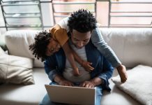 Father using the laptop trying to work while son is on his back at home, childrearing