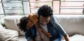 Father using the laptop trying to work while son is on his back at home, childrearing