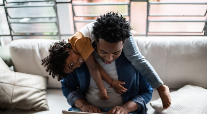 Father using the laptop trying to work while son is on his back at home, childrearing