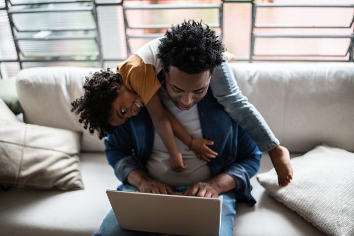 Father using the laptop trying to work while son is on his back at home, childrearing