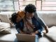 Father using the laptop trying to work while son is on his back at home, childrearing