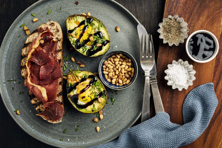 Overhead view of a dish of baked eggs with avocado, crispy serrano ham, toasted sourdough bread. Recipe, cut the avocado in half and remove the stone, use small sized eggs, discard some of the egg white if desired and pour the remaining yolk and white into the hole left by the avocado stone, season with salt and pepper and some chives and or some chilli flakes and in this case some roasted pine nuts. Bake for around 10-12 minutes at 200 degrees until the egg whites have set but the yolks are still runny. Serve with toasted bread and balsamic vinegar, delicious.