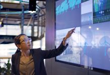 Stock photograph of a young Asian woman conducting a seminar / lecture with the aid of a large screen. The screen is displaying data & designs concerning low carbon electricity production with solar panels & wind turbines. These are juxtaposed with an image of conventional fossil fuel oil production.