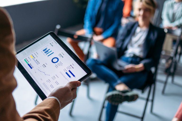 Close up photo of man hand holding tablet while group of business people listening to his analysis.