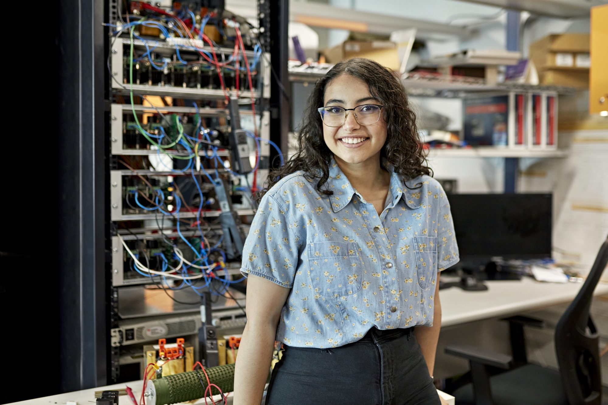 Waist-up view of casually dressed woman standing in electrical engineering lab and smiling at camera. Property release attached.