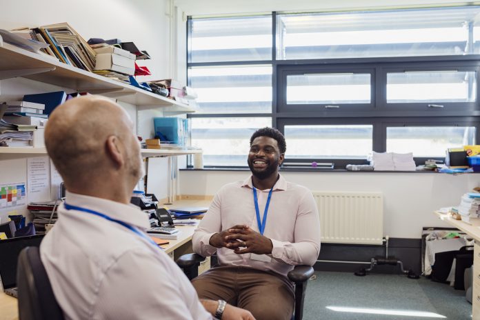 Two teachers sitting in an office together in the school that they work at in Gateshead, North East England. They are discussing lesson plans while smiling.