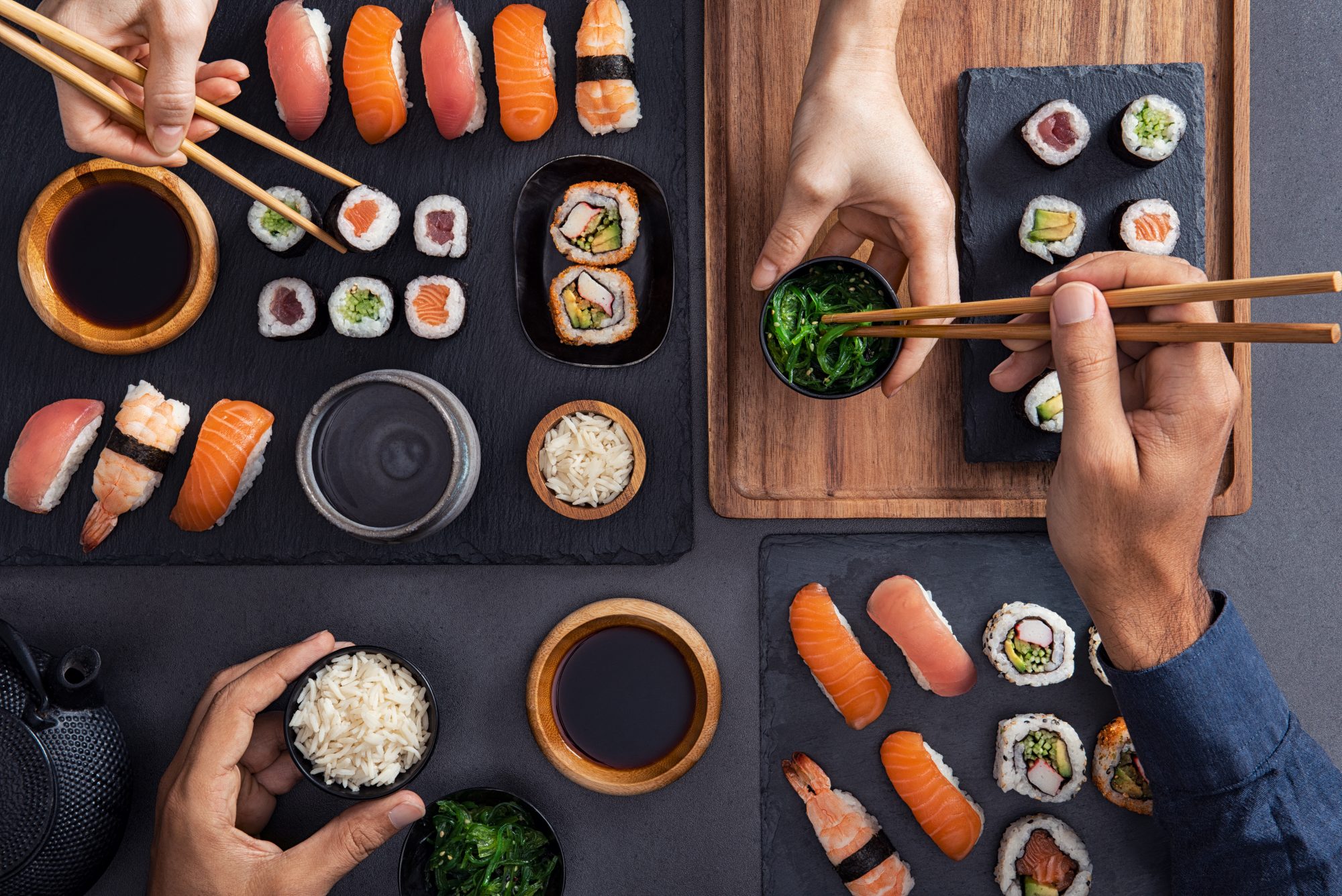 Top view of couple hands eating sushi food at japanese restaurant. High angle view of woman hand serving seaweed in little bowl with sesame to man while holding hosomaki with chopsticks. Couple eating and sharing sushi roll, maki, nigiri, uramaki.