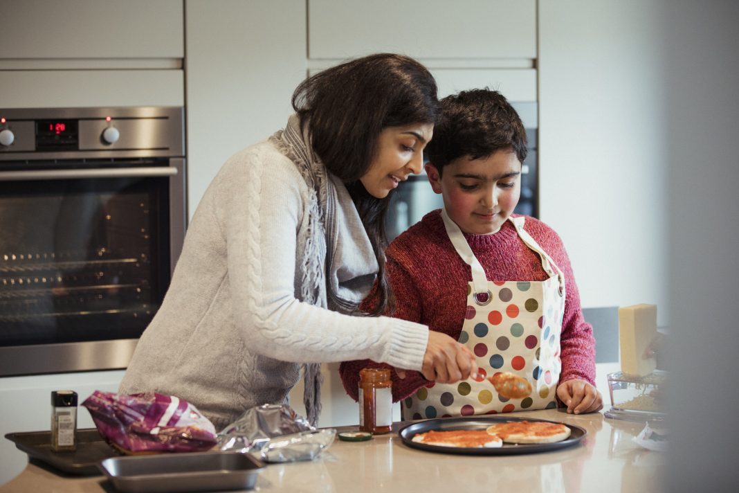 A mother is helping her autistic son spread the tomato sauce over the pizza dough in preparation for dinner.