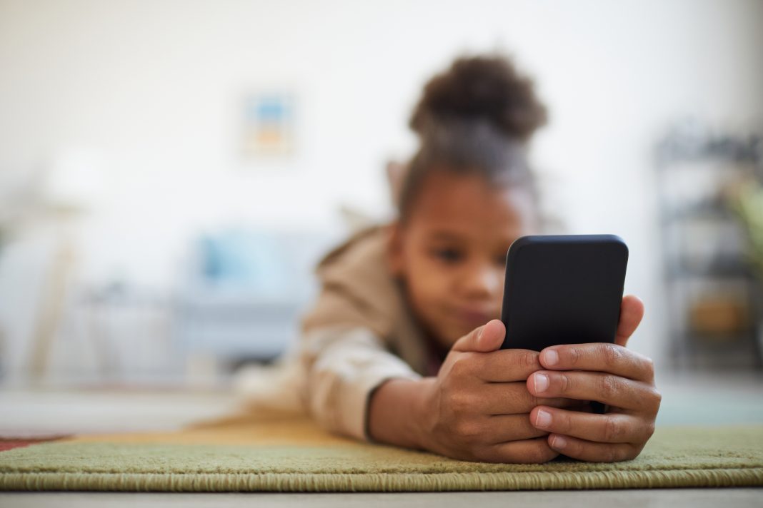 Close up of at cute African-American girl holding smartphone while lying on floor in cozy home interior, copy space