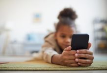 Close up of at cute African-American girl holding smartphone while lying on floor in cozy home interior, copy space