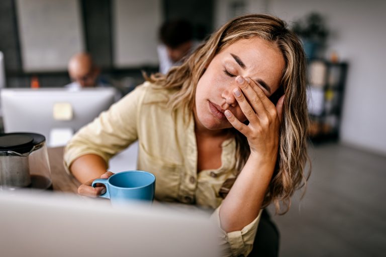 Tired and exhausted young female entrepreneur drinking coffee and rubbing eyes while feeling stressed and worried in coworking space