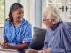 Senior Man At Home Talking To Female Nurse Or Care Worker In Uniform Making Notes In Folder, Social prescribing & building for health in the NHS