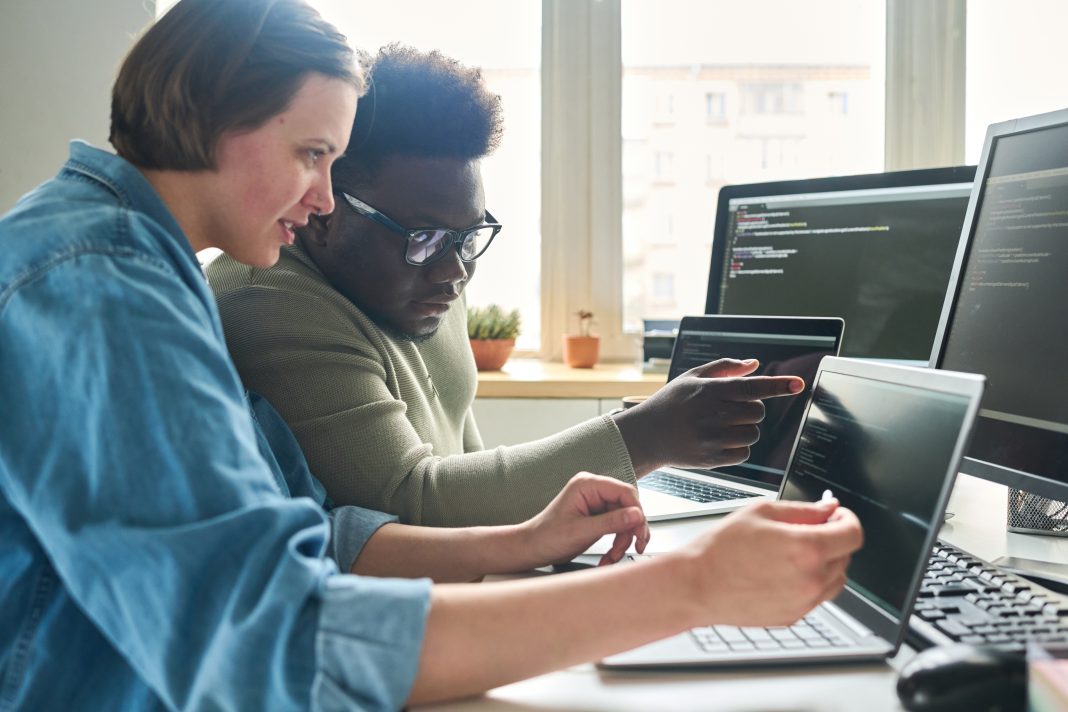 Female programmer pointing at laptop with software and consulting with colleague about computer codes