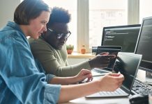 Female programmer pointing at laptop with software and consulting with colleague about computer codes