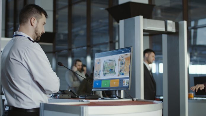Man in uniform standing at counter at checking point and watching at monitor with x-ray of luggage