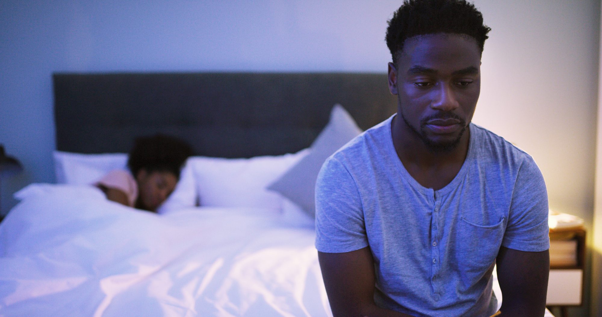 Shot of a concerned looking young man sitting on a bed while his wife sleeps in the background