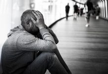 Monochrome image depicting a side view of a young homeless man sitting alone in a futuristic illuminated subway tunnel in the city. The man is clearly sad, and has an unkempt beard. He has his head in his hands in a gesture of despair. He sits with his back to the wall of the tunnel while the diminishing perspective of the subway tunnel recedes into the distance. People are defocused in the distance as they walk past, ignoring the young homeless man. Room for copy space.