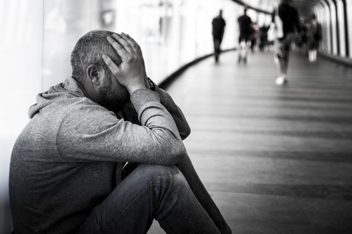 Monochrome image depicting a side view of a young homeless man sitting alone in a futuristic illuminated subway tunnel in the city. The man is clearly sad, and has an unkempt beard. He has his head in his hands in a gesture of despair. He sits with his back to the wall of the tunnel while the diminishing perspective of the subway tunnel recedes into the distance. People are defocused in the distance as they walk past, ignoring the young homeless man. Room for copy space.