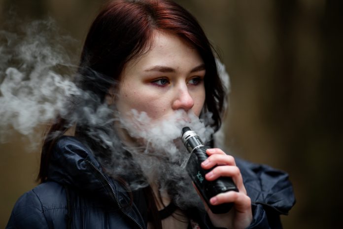 Vape teenager. Young pretty caucasian brunette girl smoking an electronic cigarette on the street in the spring. Deadly bad habit.