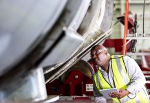 African-American man working in a manufacturing facility specializing in metal fabrication. He is wearing safety glasses and a reflective vest, holding a digital tablet as he inspects a large metal object.