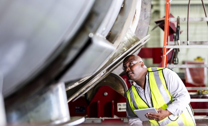 African-American man working in a manufacturing facility specializing in metal fabrication. He is wearing safety glasses and a reflective vest, holding a digital tablet as he inspects a large metal object.