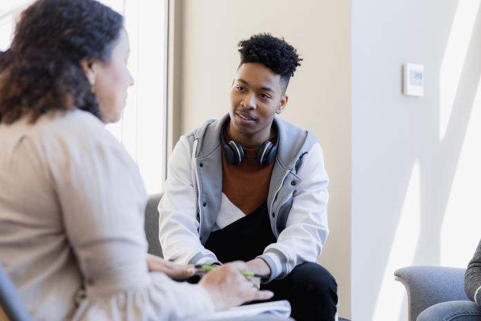 In therapy, the mid adult female counselor talks to the attentive young man and his unseen sister.