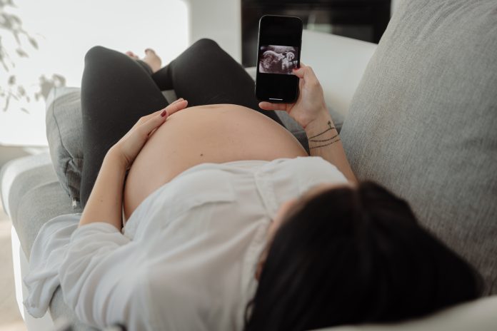 Pregnant woman lying on sofa and looking at ultrasound scans of her unborn baby at home