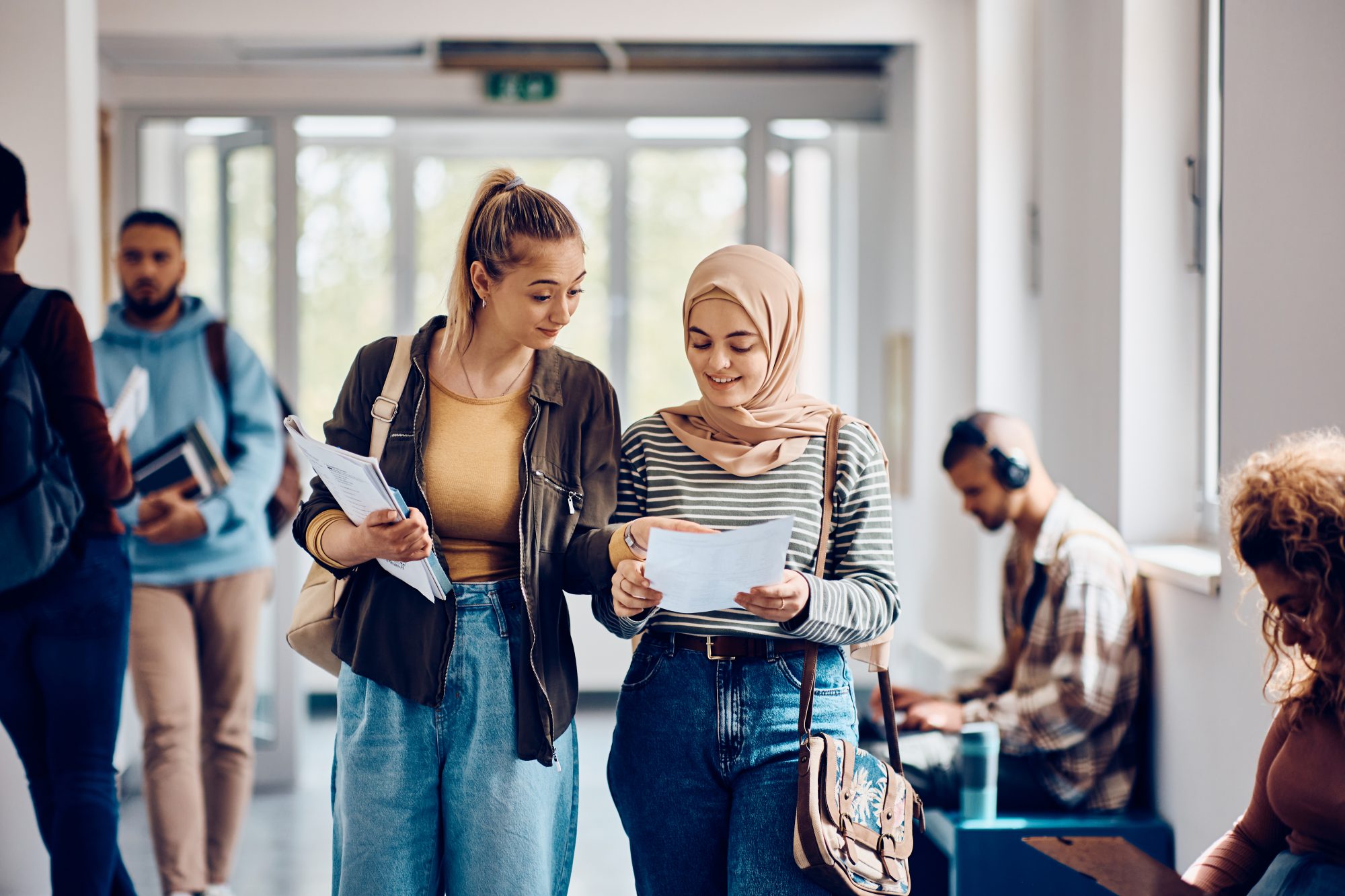 Happy Muslim student and her friend reading their exam results while walking through university hallway.