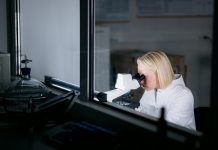 Blonde female scientist wearing a lab coat, working in her laboratory and conducting diverse experiments, using a microscope and looking through it