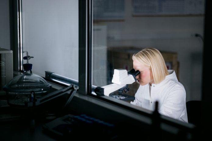 Blonde female scientist wearing a lab coat, working in her laboratory and conducting diverse experiments, using a microscope and looking through it