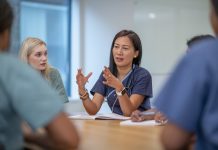 A small group of Nurse sit at a boardroom table as they meet to discuss patient cases. They are each dressed professionally in scrubs and have stethoscopes around their necks as they discuss plans of care for each case.