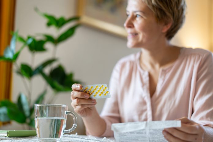 A smiling woman is holding a blister packet or HRT pills and the leaflet, with a drink of water ready on the table sitting at home