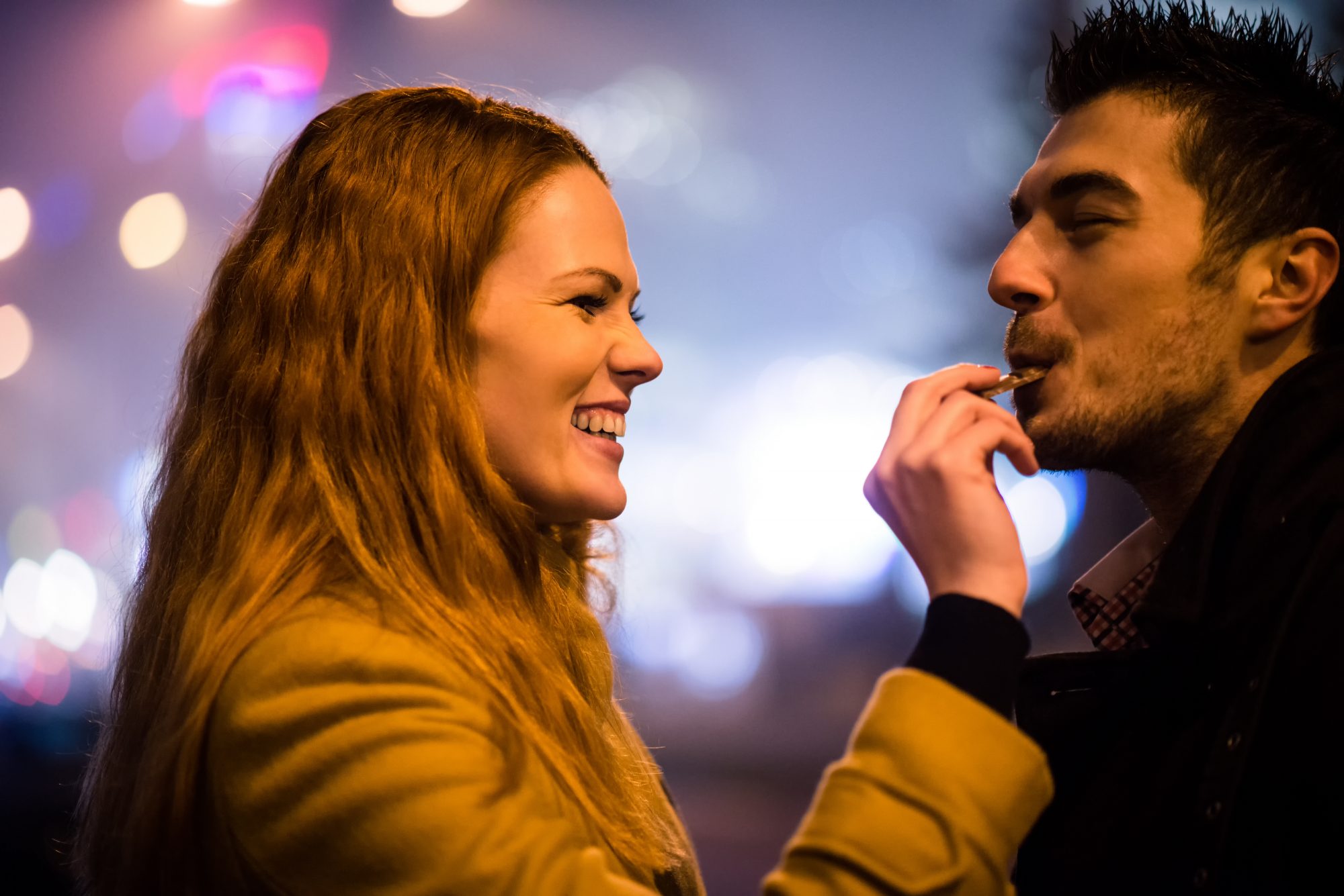 Couple on date - woman feeds her boyfriend with chocolate in street at winter night