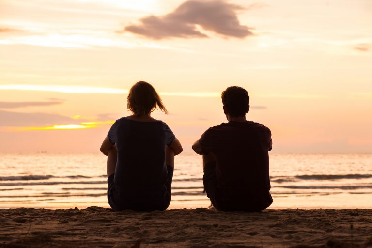 Friends sitting together on the beach and watching sunset, friendship concept