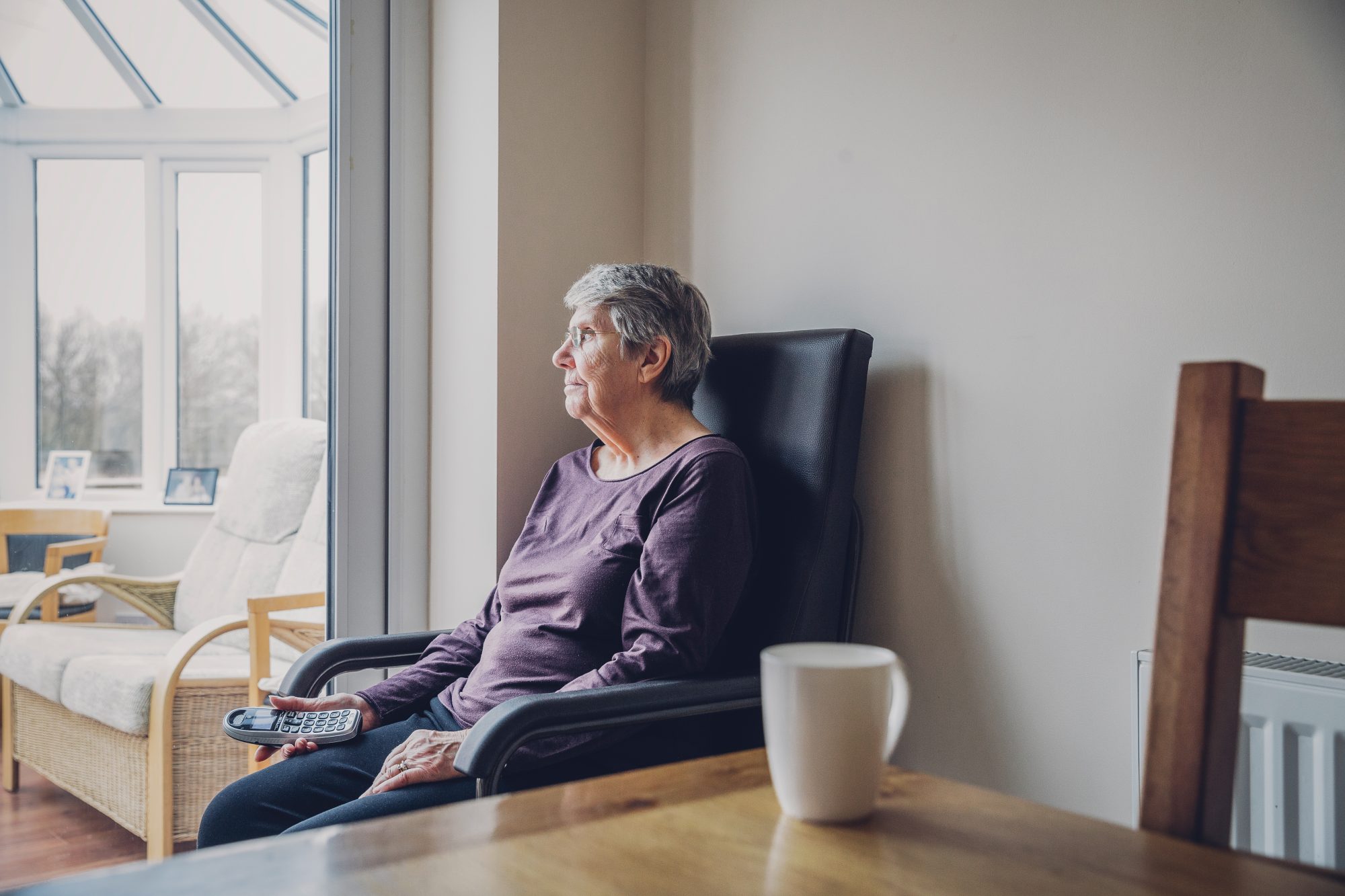 Senior woman sitting alone in her kitchen. She is looking out into her conservatory while holding a home telephone in her hand. Serious expression