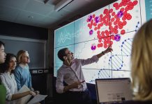 Group of business professionals in a dark room standing in front of a large data display screen with information.