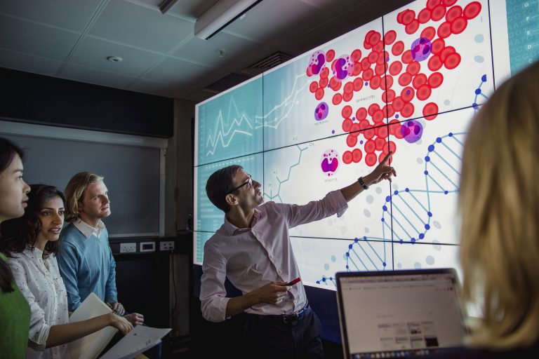 Group of business professionals in a dark room standing in front of a large data display screen with information.