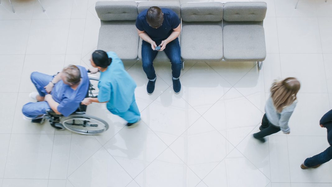 High Angle Shot in the Hospital Lobby, Young Man Waits for Results while Sitting and Using Mobile Phone, Doctors, Nurses and Patients Walk Past Him. Clean, New Hospital.